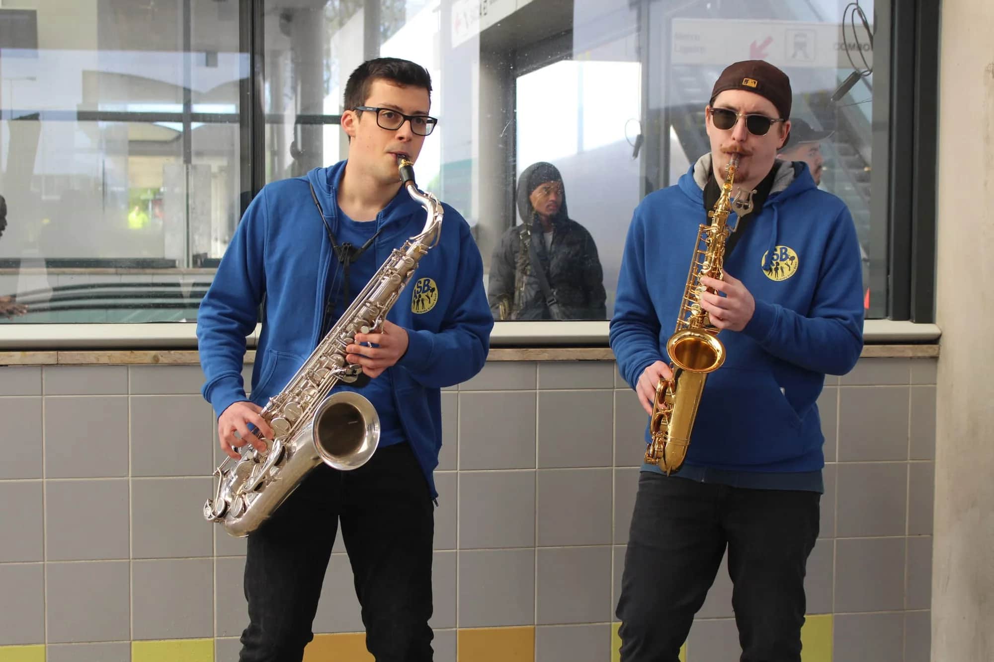 Dois saxofonistas a tocar numa estação, com uma pessoa a olhar da janela